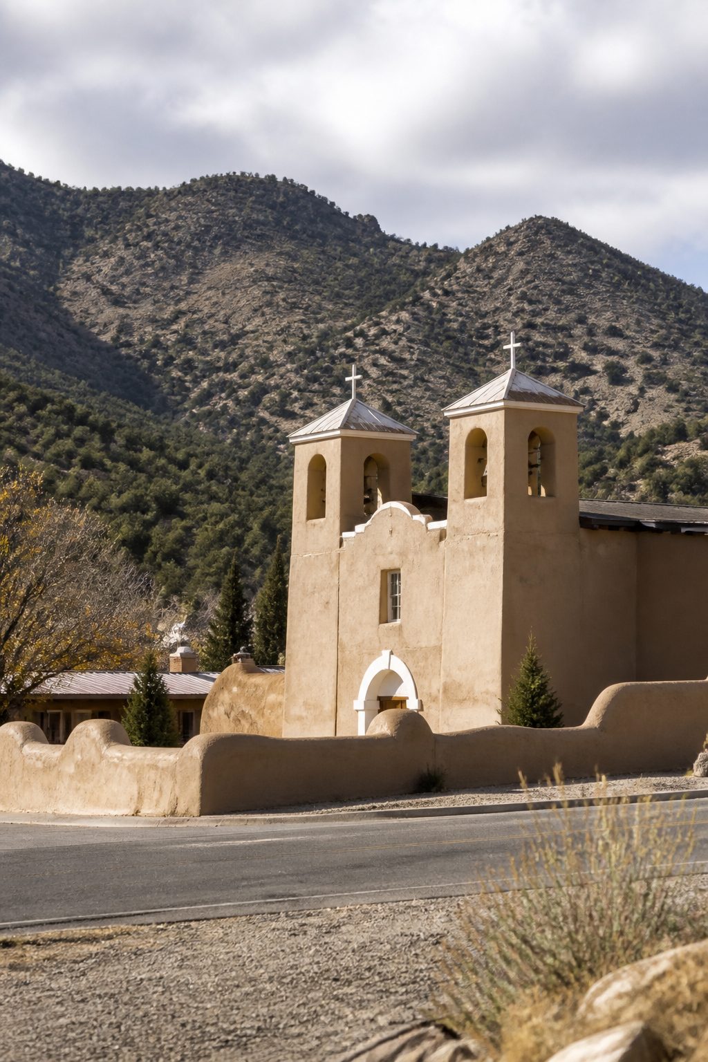 An adobe church with twin bell towers and a low mud-plastered wall at the centre of a small mountain village, with pinon-covered hills rising behind it