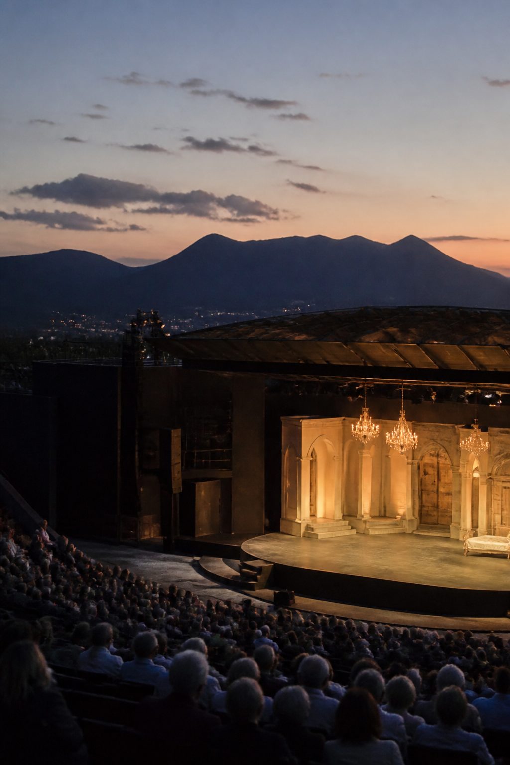 An open-sided opera theatre at dusk with the stage lit and the dark outline of distant mountains visible behind it