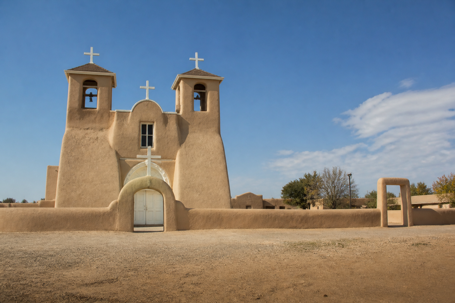 A twin-towered adobe mission church with a low white cross above the door and a walled earthen churchyard, standing empty at the edge of a bare dirt plaza under a wide blue sky