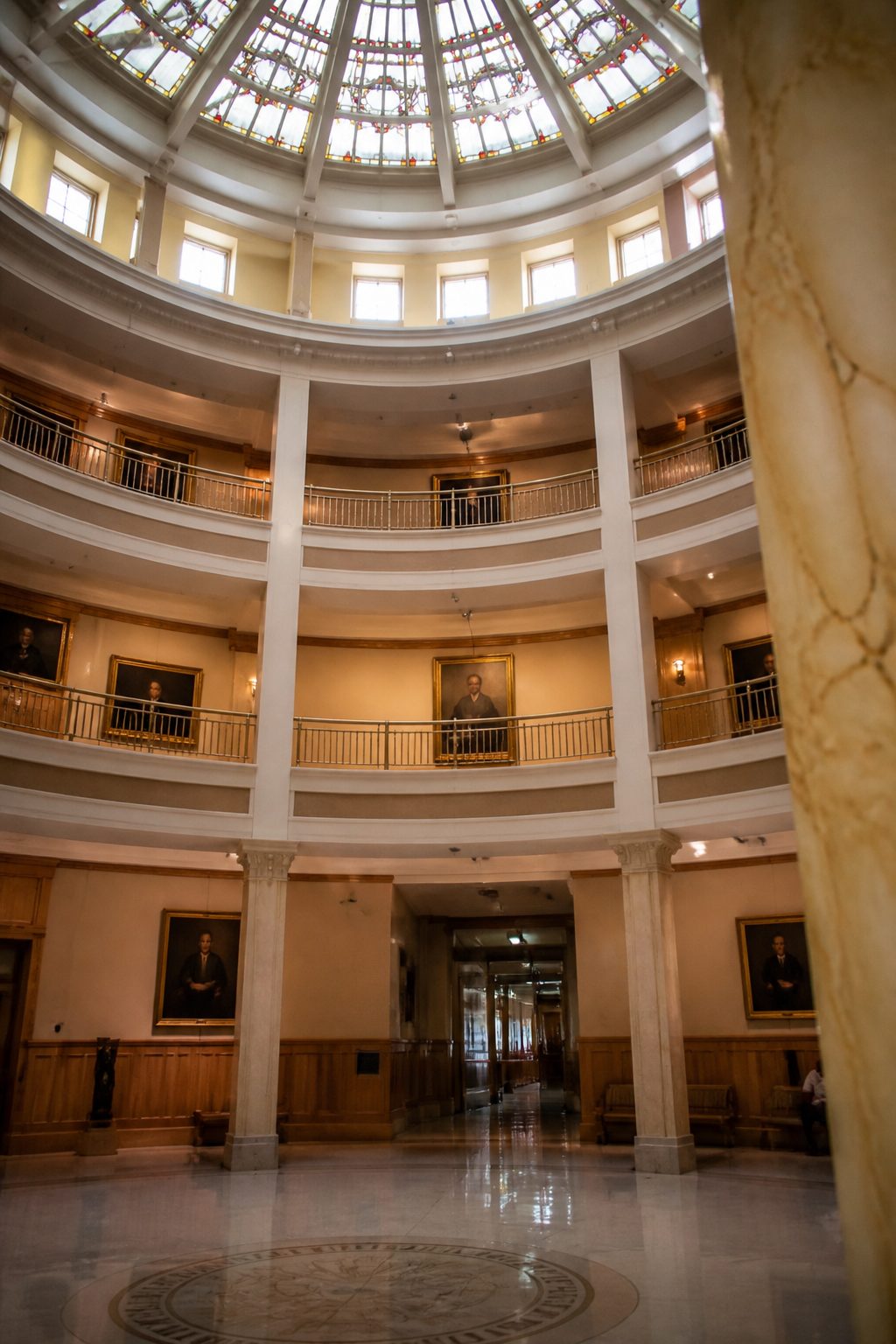 The circular rotunda of the New Mexico State Capitol seen from the ground floor, with tiered balconies on each level and a stained glass ceiling above