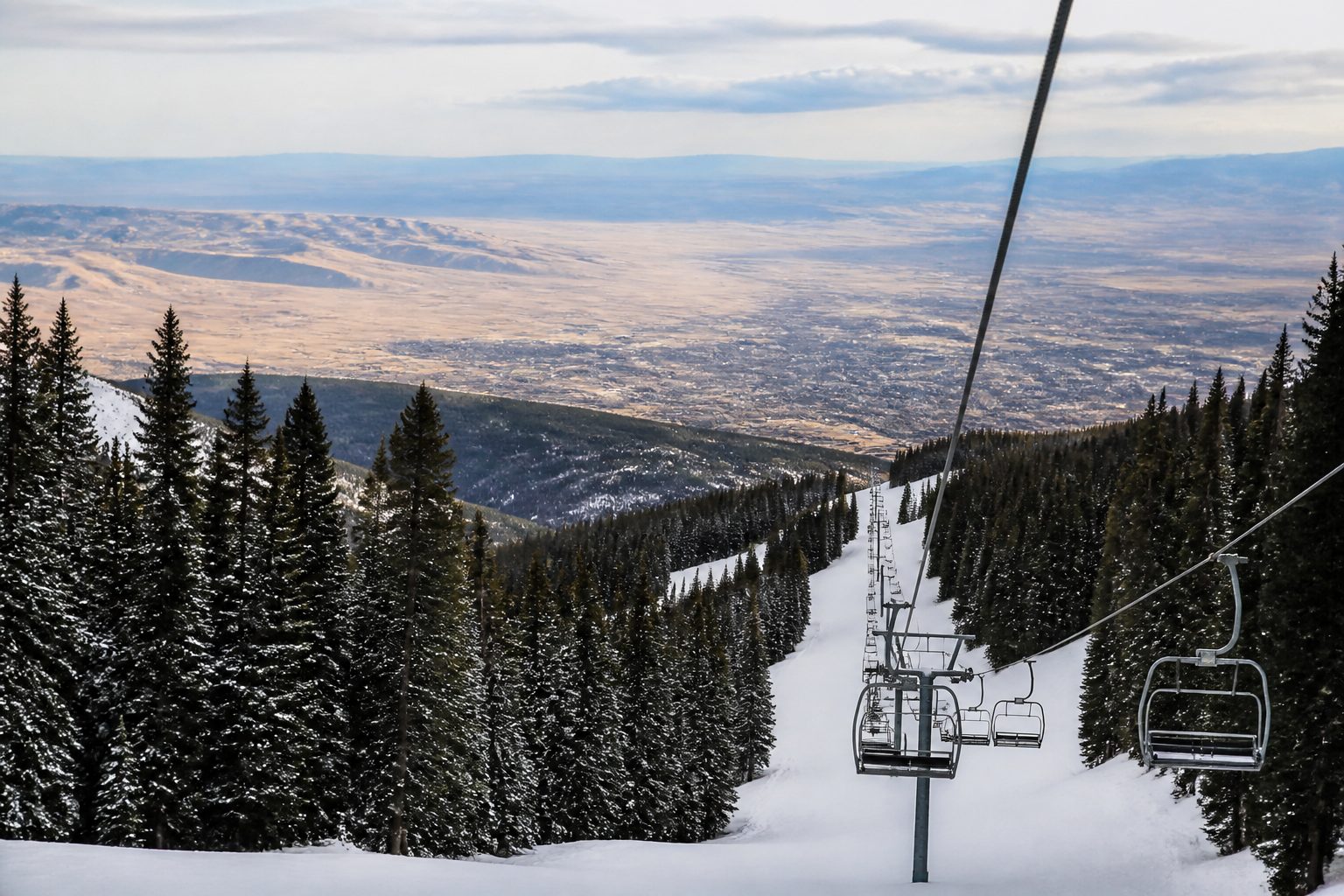Snow-covered pistes cut between dark spruce and fir on a north-facing mountainside, a chairlift line running up the slope and a wide brown valley visible far below